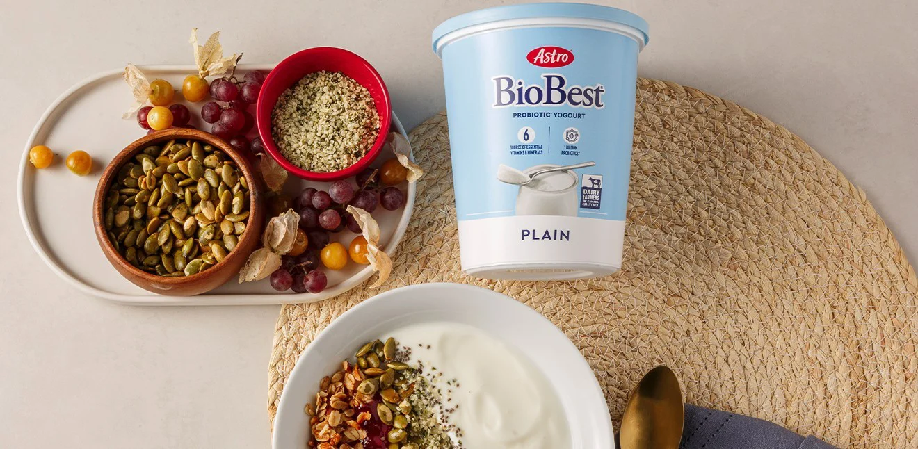 Astro Biobest Yogurt tub on a table with various grains, assorted frut and a bowl of yogurt topped with seeds and grains.