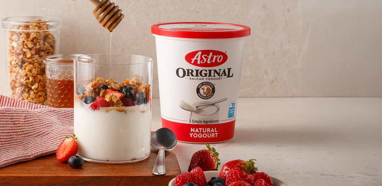 Astro Plain 6% yogurt tub on a table with yogurt in a glass with honey, fruit and grains
